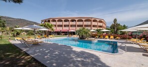 Ein Schwimmbecken mit Liegestühlen und Sonnenschirmen, umgeben von einem Gebäude mit Terrakotta-Dach und mehreren Balkonen.