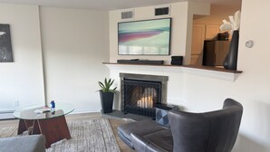 A living room with a fireplace, a flat-screen TV mounted above it, a modern armchair, a glass coffee table, and a potted plant. - 2 Bedroom Condo -Views of West Point and Hudson River, steps from Metro North! (Garrison)