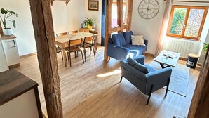 A rustic interior with exposed wooden beams, a dining area with a wooden table and chairs, a blue sofa, and a window with a view of greenery. - La P'tite Maison - Authentic stay on the Wine Route (Orschwihr)