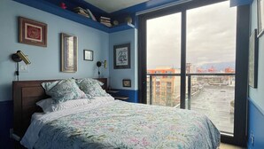 A bedroom with a bed, ceiling fan, and large window with a view of buildings.
