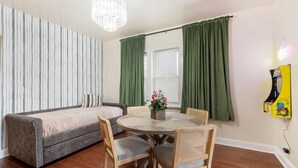 Una habitación con un sofá gris, una mesa redonda con sillas, una araña y cortinas verdes.