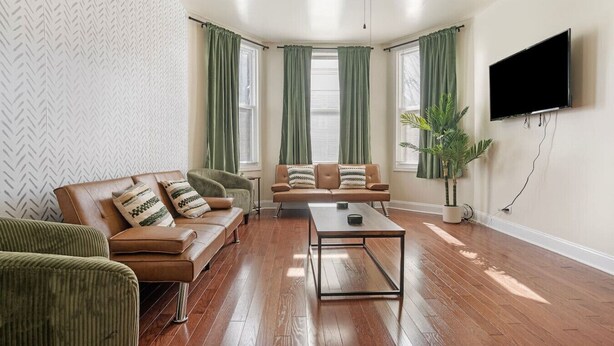Una sala de estar con un sofá, sillón, mesa de centro y televisor montado en la pared.