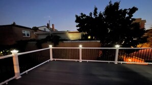 A rooftop deck with a white railing, a small light fixture, and a view of surrounding buildings and trees.