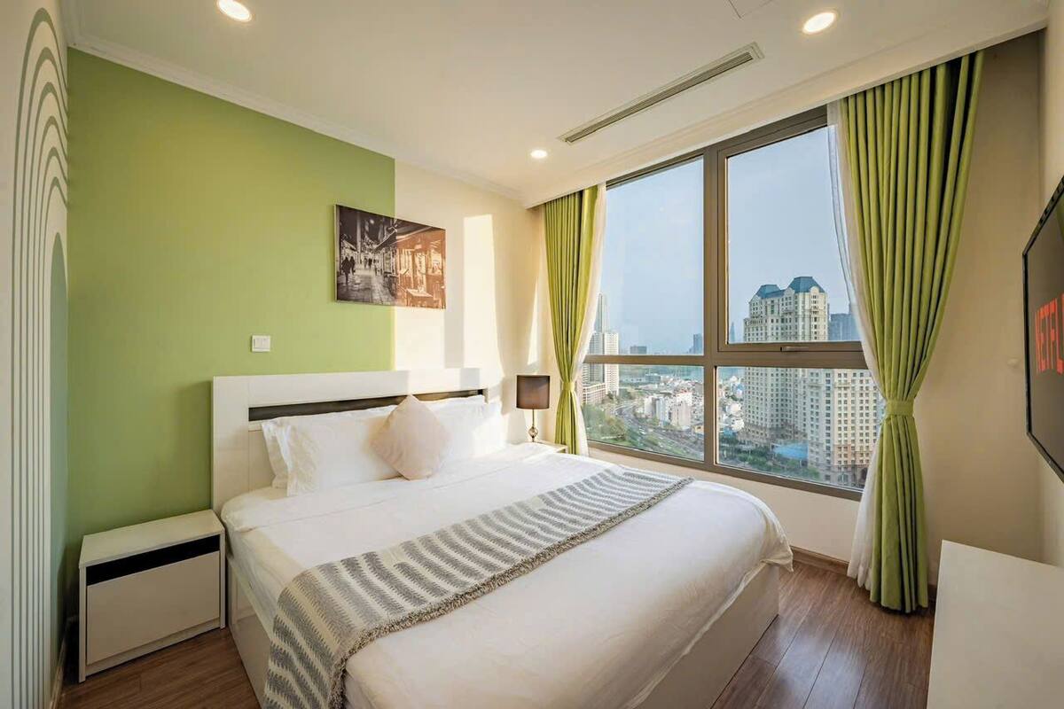 Ein Hotelzimmer mit einem großen Bett, einem Nachttisch, einem Fenster mit Vorhängen und Blick auf die Stadt.