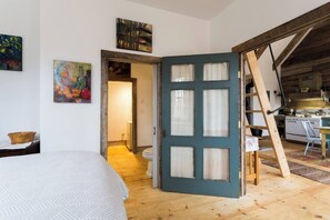 Une pièce comprenant un lit, un plancher en bois, un coin cuisine et une porte donnant sur une autre pièce.