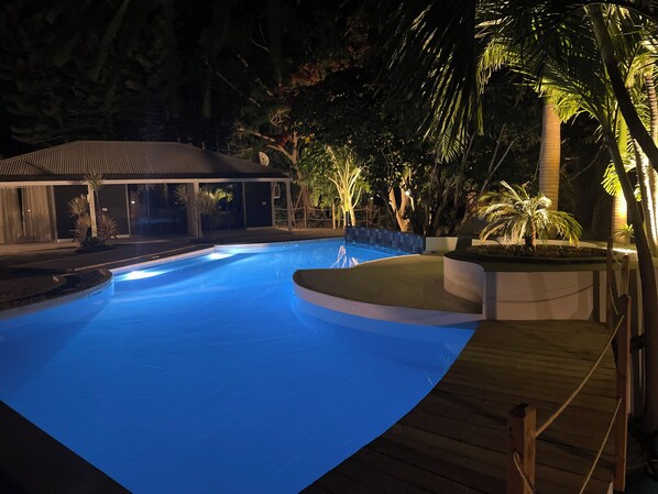 A swimming pool with a wooden deck, surrounded by palm trees and a covered area. - Résidences de la Métisse (Saint-François)