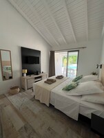 A spacious bedroom with a large bed, a flat-screen TV mounted on the wall, a wooden dresser, and a large window with a view of greenery outside.