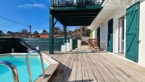 A wooden deck with a pool, outdoor furniture, and a building with green shutters. - Spacious house with garden and pool (Arcachon)