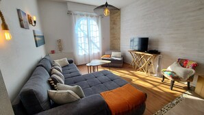 A modern living room with a sectional sofa, a small table, a TV on a wooden stand, and a window with curtains. - Spacious house with garden and pool (Arcachon)