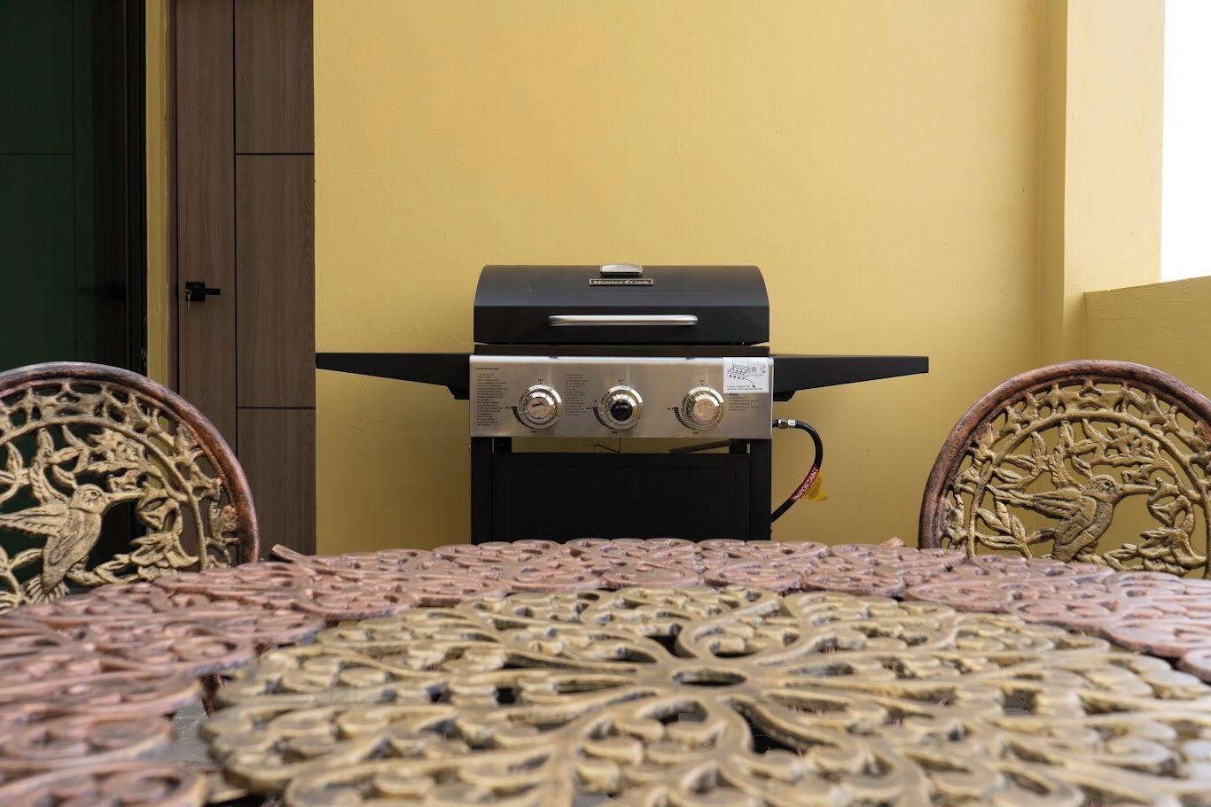 A small outdoor grill area with a table and chairs, a yellow wall, and a door in the background.