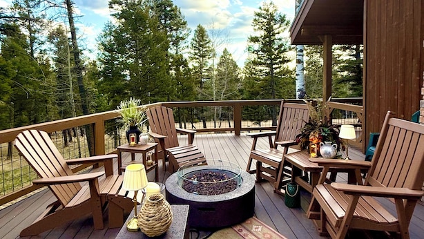 A wooden deck with Adirondack chairs, a glass fire pit, and a table with a lamp and decorative items. - Grinning Bear Lodge Luxury Cabin, Hot Tub, Massive Deck, Sleeps 14, Game Loft (Angel Fire)
