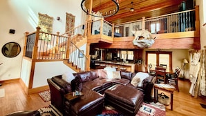 A spacious living area with a leather sofa, a dining table, and a staircase leading to an upper level. - Grinning Bear Lodge Luxury Cabin, Hot Tub, Massive Deck, Sleeps 14, Game Loft (Angel Fire)