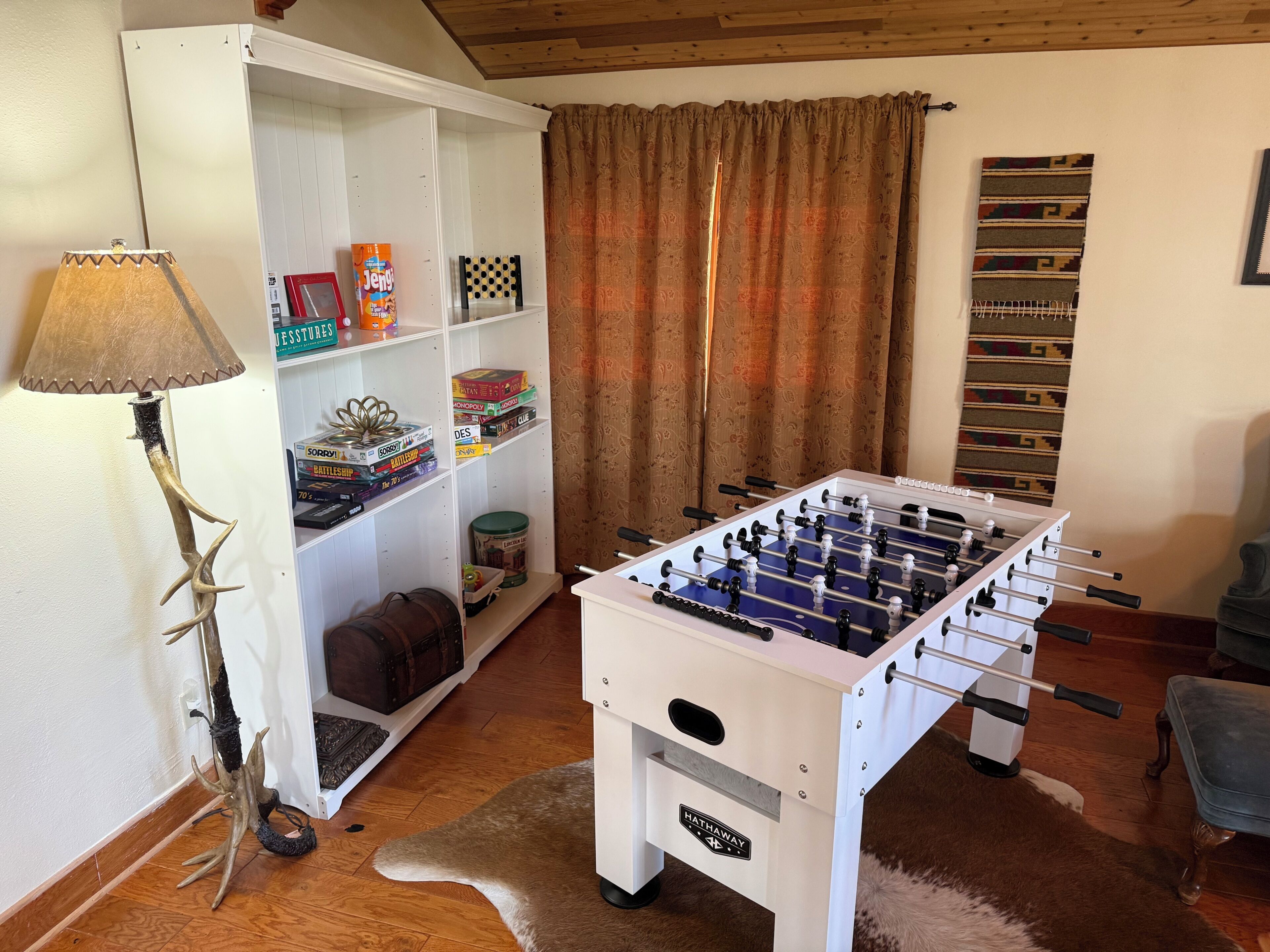 Una habitació amb taula de futbolí, una estanteria plena de jocs de taula, una làmpada i una catifa de pell.
