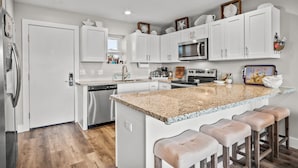 A modern kitchen with white cabinetry, granite countertops, and a central island with bar stools. - Stay in this 3BR/2BA condo you may never want to leave! (Port Aransas)