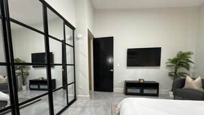 Ein modernes Schlafzimmer mit einem großen Bett, einer Glastür, die sich öffnen lässt, einem schwarzen Kleiderschrank, einem an der Wand montierten Fernseher und einer kleinen Pflanze.