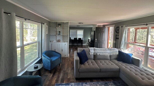 Ein geräumiges Wohnzimmer mit großem Fenster, einer Couch, einem blauen Sessel und einem Essbereich im Hintergrund.