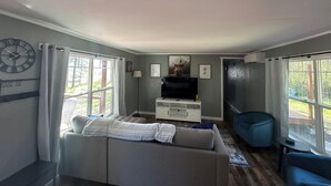 Ein Wohnzimmer mit einer Couch, einem Fernseher, einer Uhr und einem Fenster mit Vorhängen.