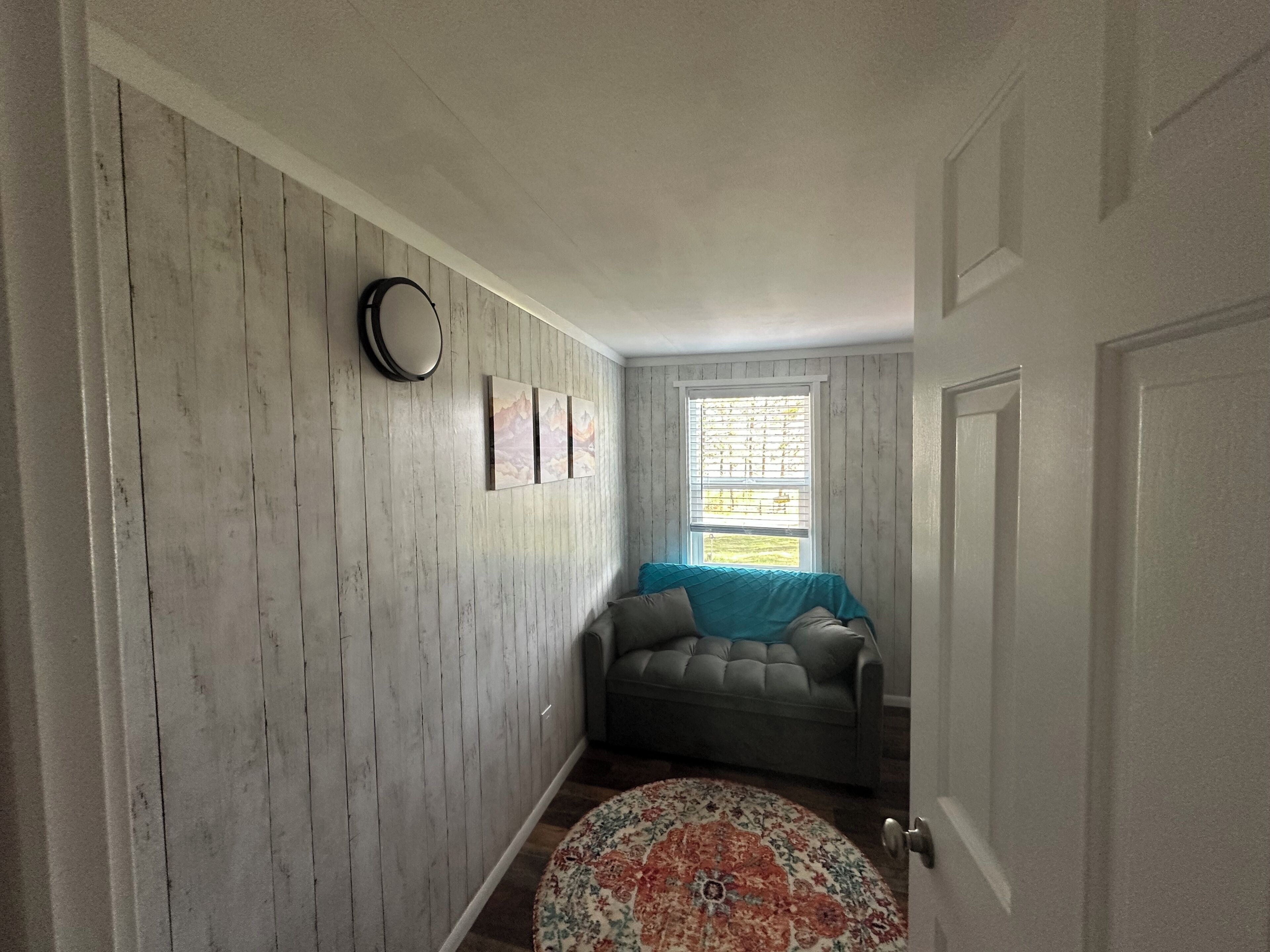 Un pasillo con pared de madera, un tapete redondo, un sofá gris y una ventana con persianas.