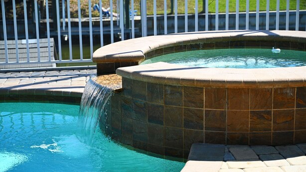 Una piscina con cascada, rodeada de baldosas de piedra y una valla de metal.