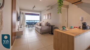 A modern living room with a sofa, a coffee table, and a dining area with chairs. There is a large window providing natural light and a view of the outdoors. - Dream stay just 250m from Praia do Farol (Salvador)