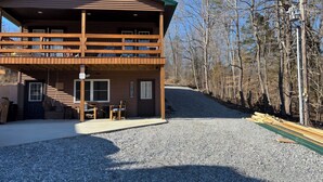 A two-story log cabin with a covered porch, a gravel driveway, and a forested backdrop.