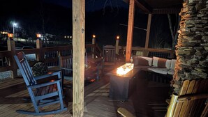 A wooden deck with a stone wall, a wooden rocking chair, a table with a lit candle, and a stone fireplace.