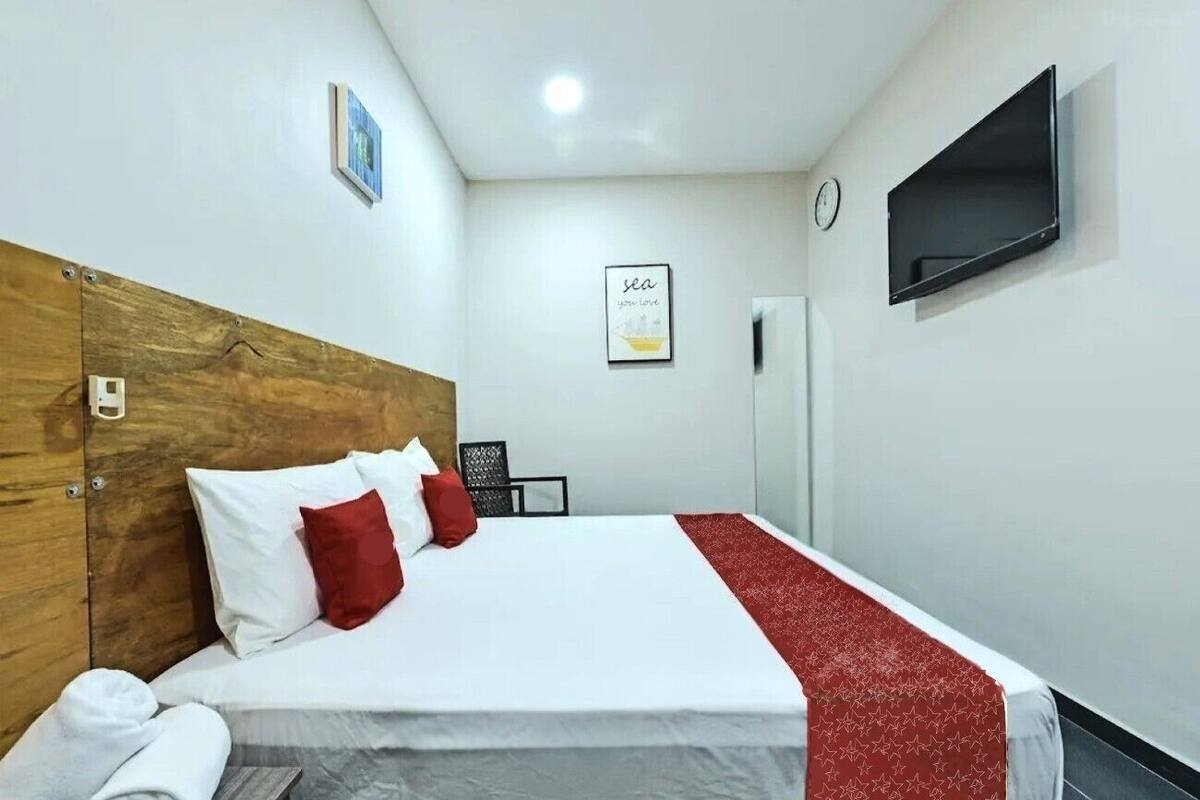 Une chambre d’hôtel avec une tête de lit en bois, un lit avec une literie blanche et rouge, un plaid rouge, un téléviseur à écran plat fixé au mur et une citation inspirante encadrée accrochée au mur.