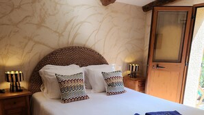 Een slaapkamer met een groot bed, een houten hoofdbord, gekleurde kussens, een nachtkastje met een lamp en een houten garderobe.