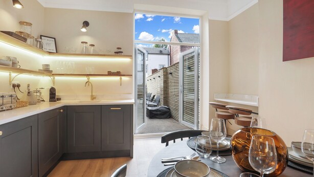 Una cocina moderna con armarios grises oscuros, una encimera de madera y un comedor con mesa dispuesta para cuatro personas. Un ventanal ofrece vistas al exterior.