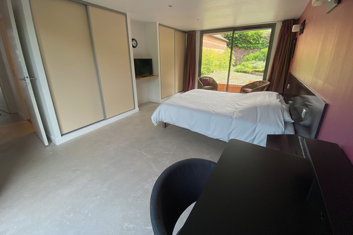 A hotel room with a bed, a desk, a chair, a TV, and a sliding door leading to a balcony with a view of greenery.