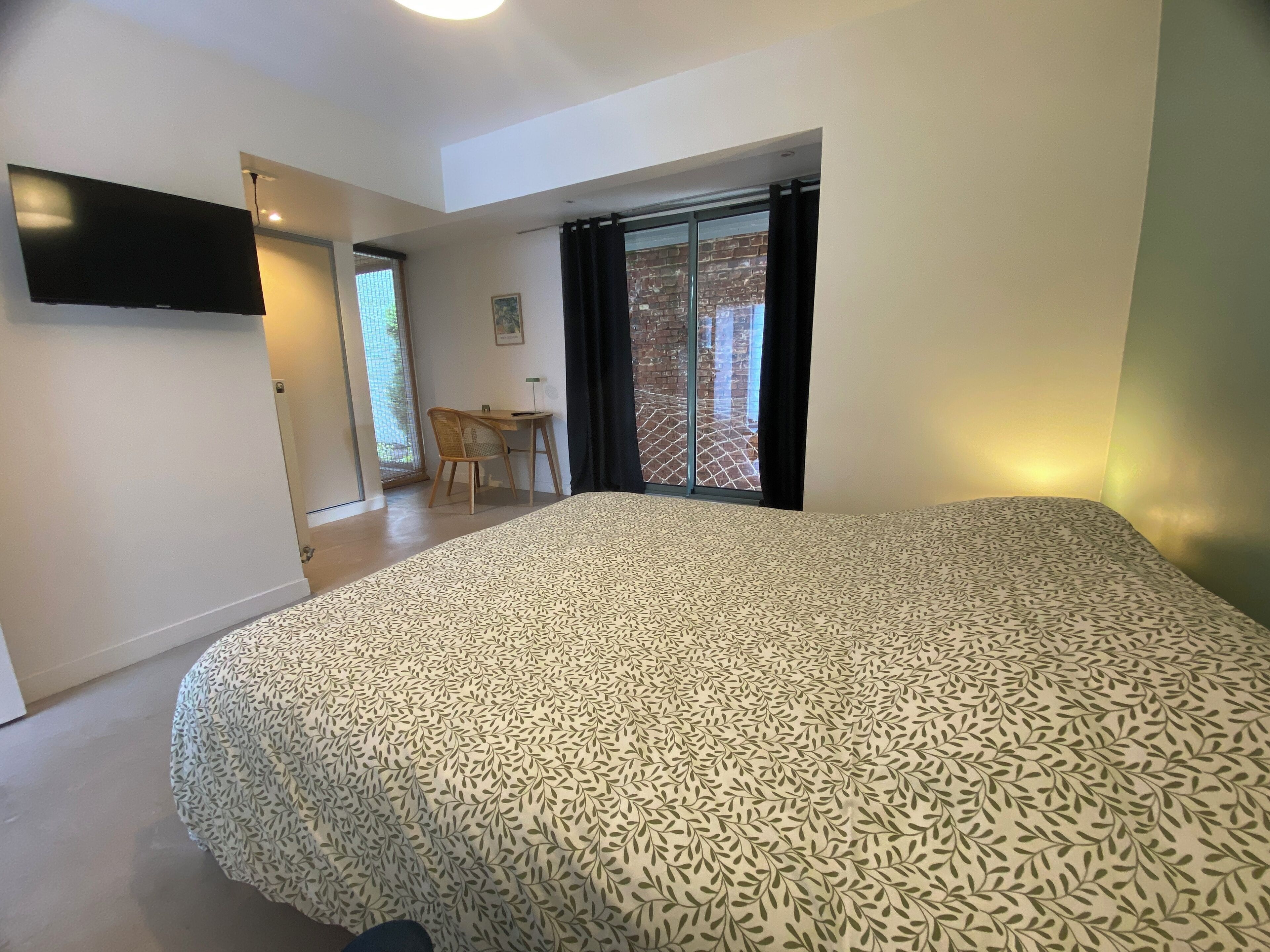 Un dormitorio con una cama grande, un escritorio y una silla. Hay una ventana que da a un muro de ladrillo y una puerta que conduce a otra habitación.