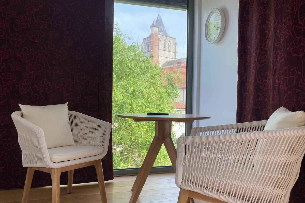 A room with two white wicker chairs, a small wooden table, and a view of a church through a window.