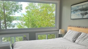 Una camera da letto con un'ampia finestra, un letto, una testiera, una lampada e un quadro appeso al muro.