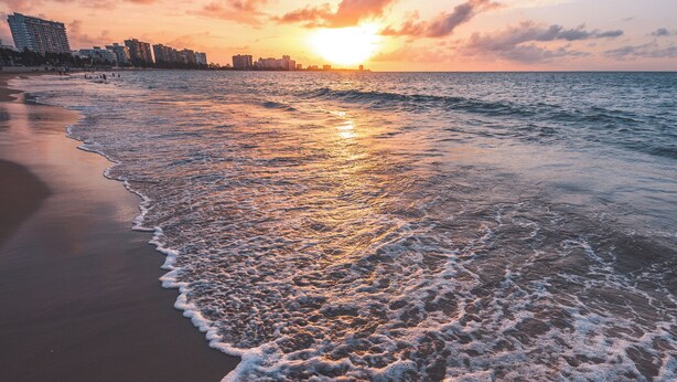 Une plage où les vagues viennent doucement lécher le rivage, un coucher de soleil en arrière-plan et, au loin, la silhouette d’une ville qui se dessine sur l’eau.