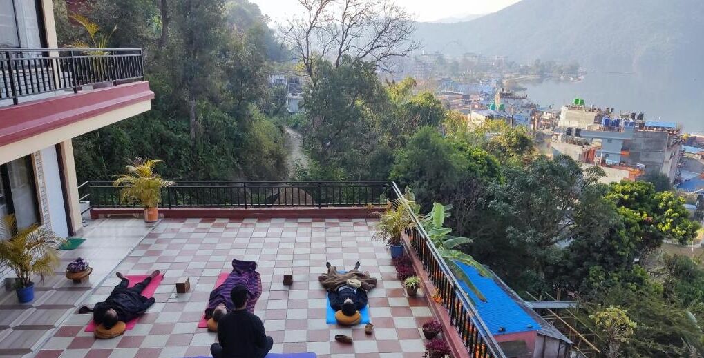 Un balcón con gente practicando yoga, rodeado de vegetación y vistas a un pueblo.