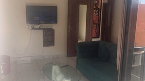 A living room with a green sofa, a glass coffee table, a flat-screen TV mounted on the wall, and a door leading to another room. - Furnished and air-conditioned 2-bedroom apartment living room (Dakar)