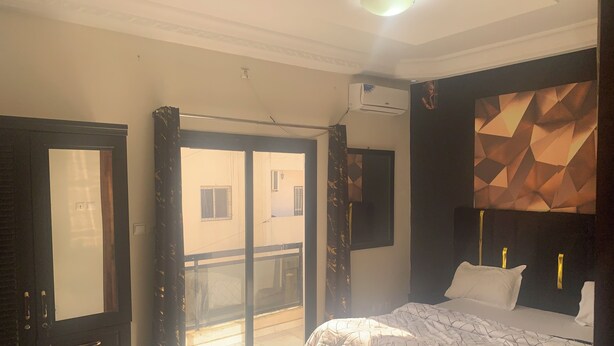 A hotel room with a bed, a sliding glass door to the balcony, a mounted air conditioner, and a geometric wall decoration. - Furnished and air-conditioned 2-bedroom apartment living room (Dakar)