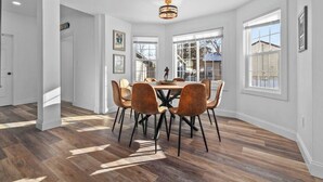 A dining area with a round table and six chairs, large windows, and a chandelier.