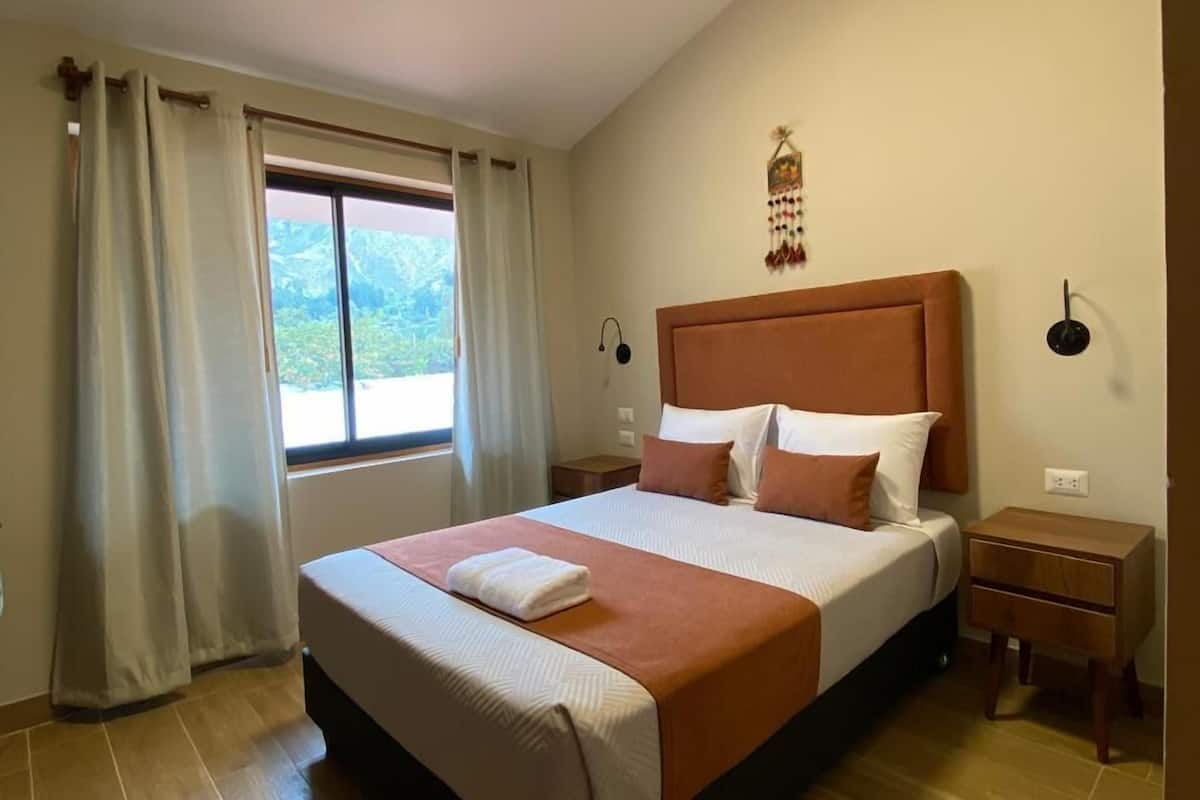 Un dormitorio ordenado con una cama grande, una mesita de noche, cortinas en una ventana y una lámpara en el techo.