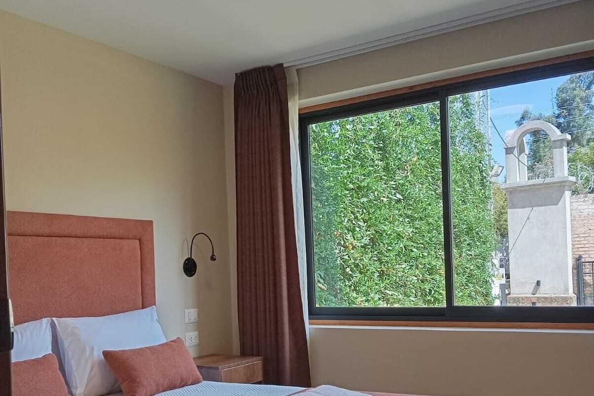 Un dormitorio con una cama grande, una mesita de noche, una lámpara de noche, un ventanal con vistas a la vegetación y una luz en el techo.