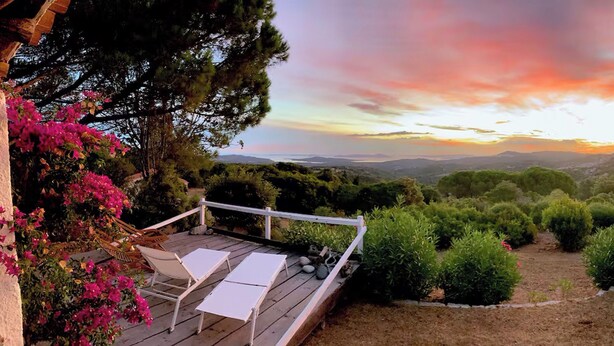 Eine Holzterrasse mit weißen Liegestühlen und Blick auf einen Sonnenuntergang über einer hügeligen Landschaft.