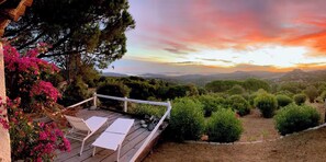 Une terrasse en bois avec des chaises longues blanches et une vue sur un coucher de soleil se déclinant sur un paysage vallonné.
