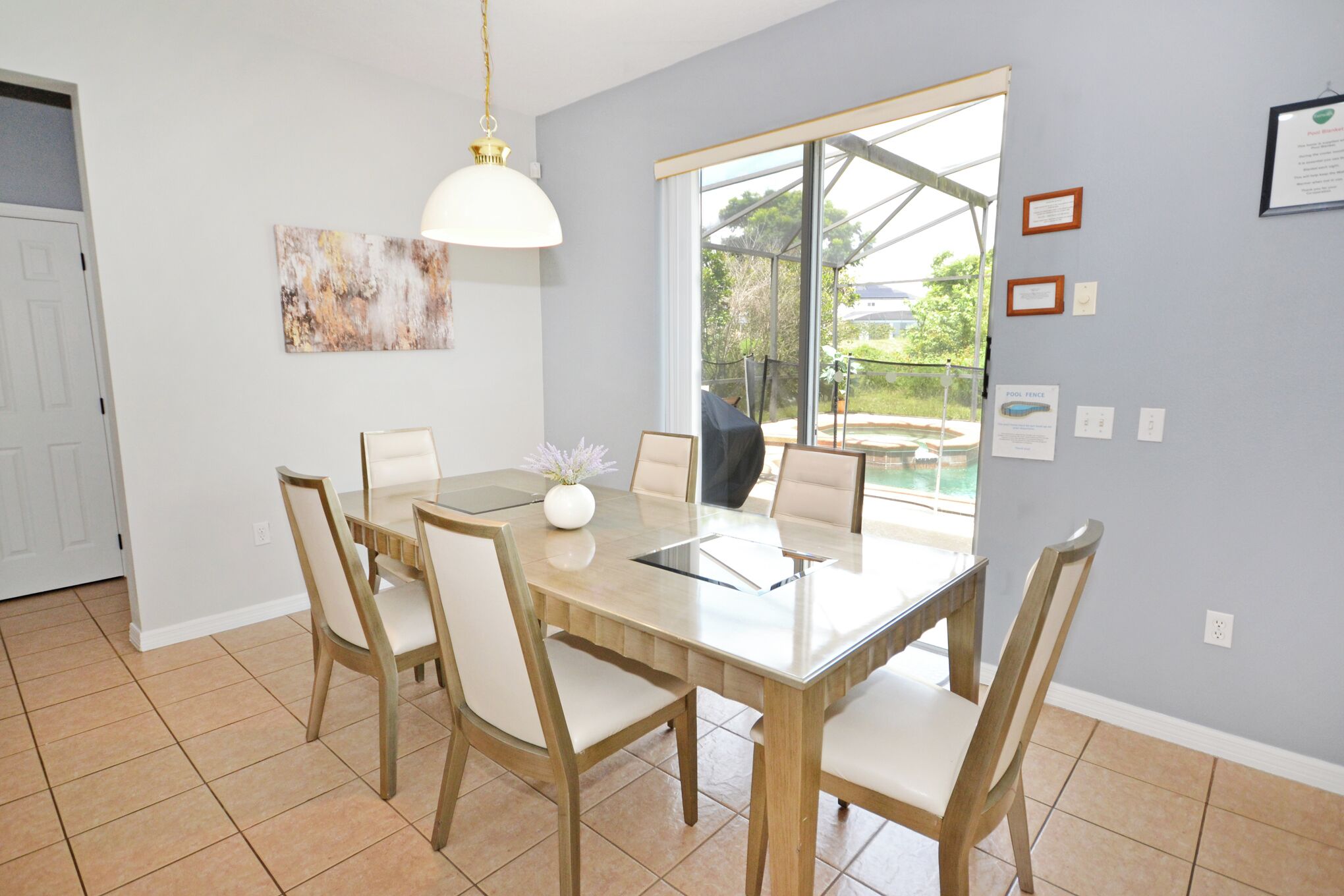 A dining area with a wooden table and chairs, a pendant light, a window with a view, and a wall with framed pictures and a certificate.