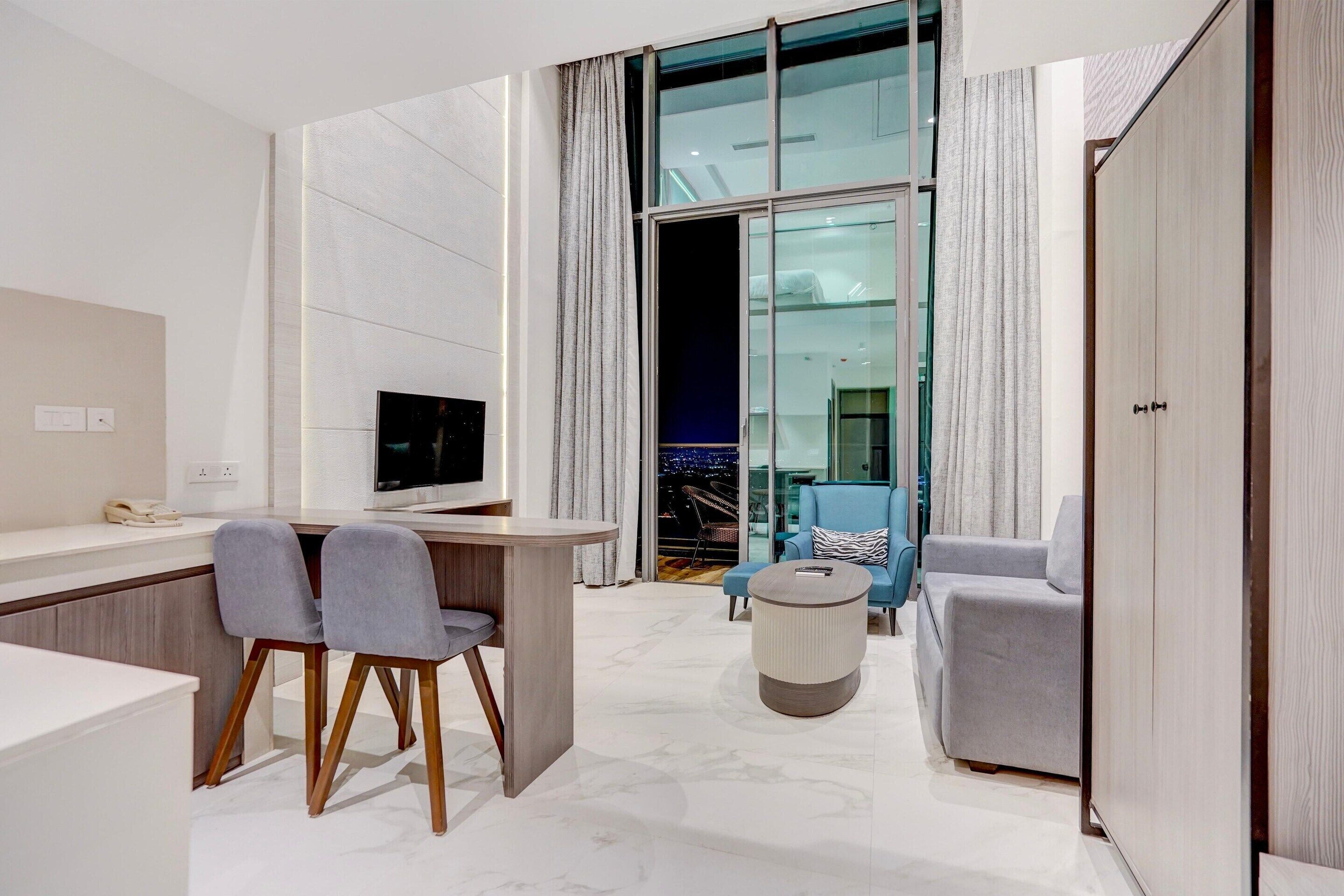 A modern hotel room with a desk, two chairs, a TV, a round coffee table, and a wardrobe.
