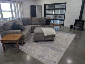 Un salon moderne avec un canapé d’angle, un petit pouf, une table basse en bois et une grande fenêtre avec des rideaux.