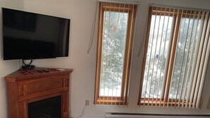 A living room with a flat-screen TV mounted on the wall, a wooden cabinet below, and a fireplace. There are two windows with blinds, and a wall-mounted heater is visible. - Family-Friendlyin QuietLaval Neighborhood (Laval)