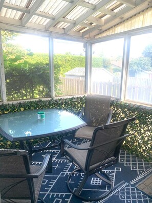Een overdekt terras met een tafel en stoelen, grote ramen en uitzicht op groen.