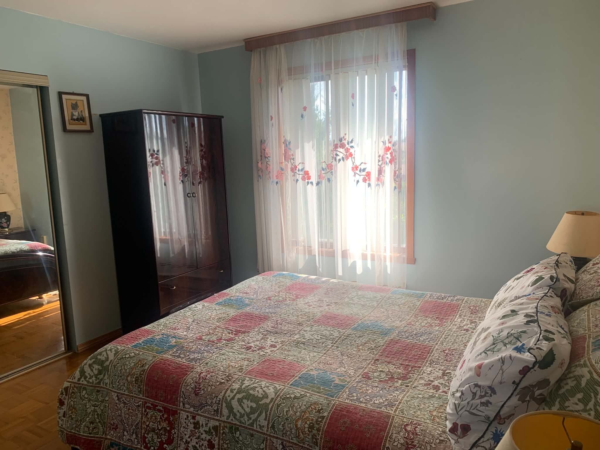 A bedroom with a bed, a wardrobe, a window with floral curtains, and a lamp.