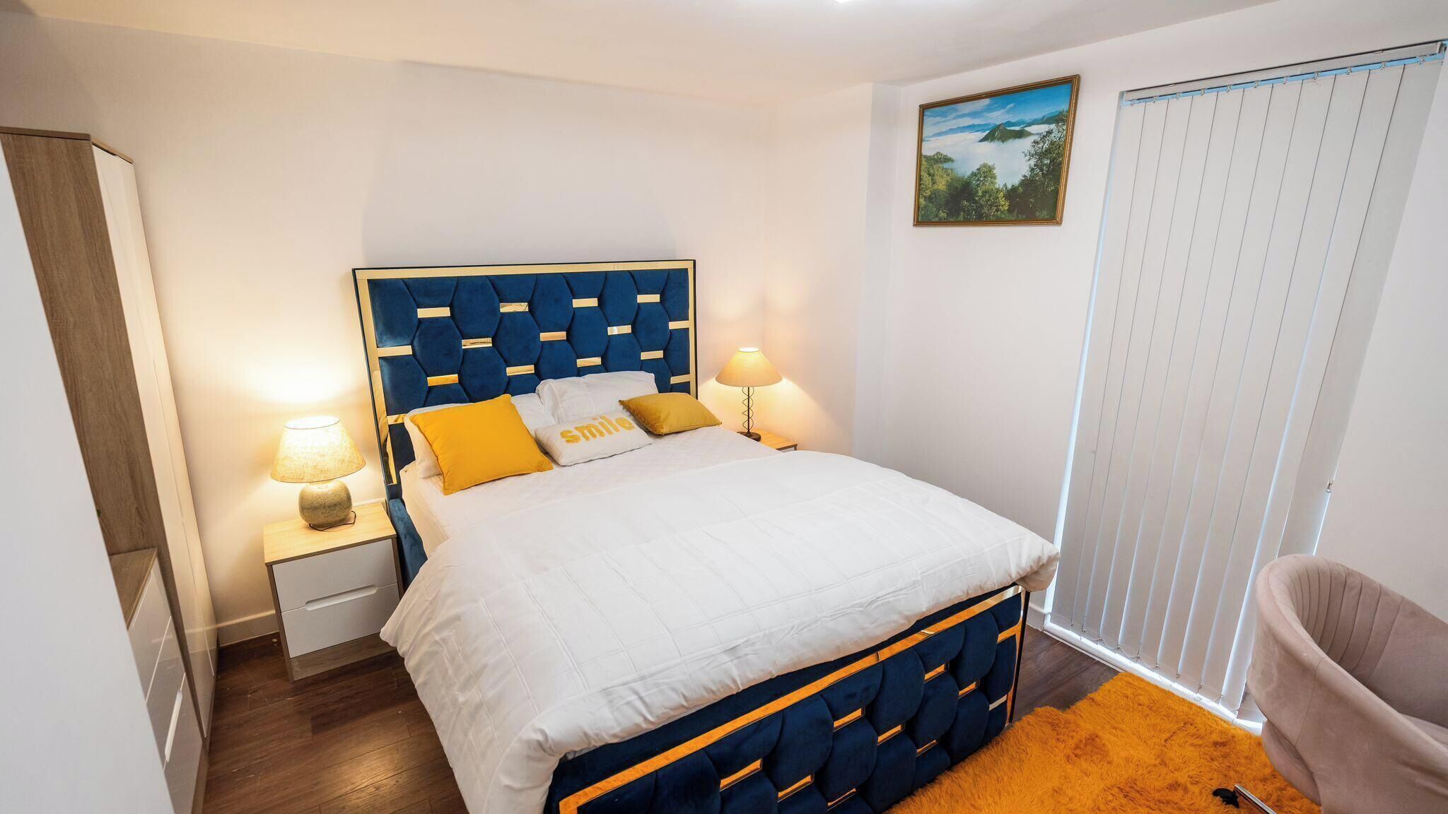 Un dormitorio con cama, lámparas de noche, mesita de noche, una silla y una ventana con persianas.
