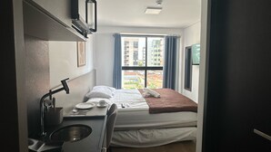 Habitación de hotel compacta con una cama individual, un escritorio pequeño con silla, una ventana con cortinas y vistas a edificios.